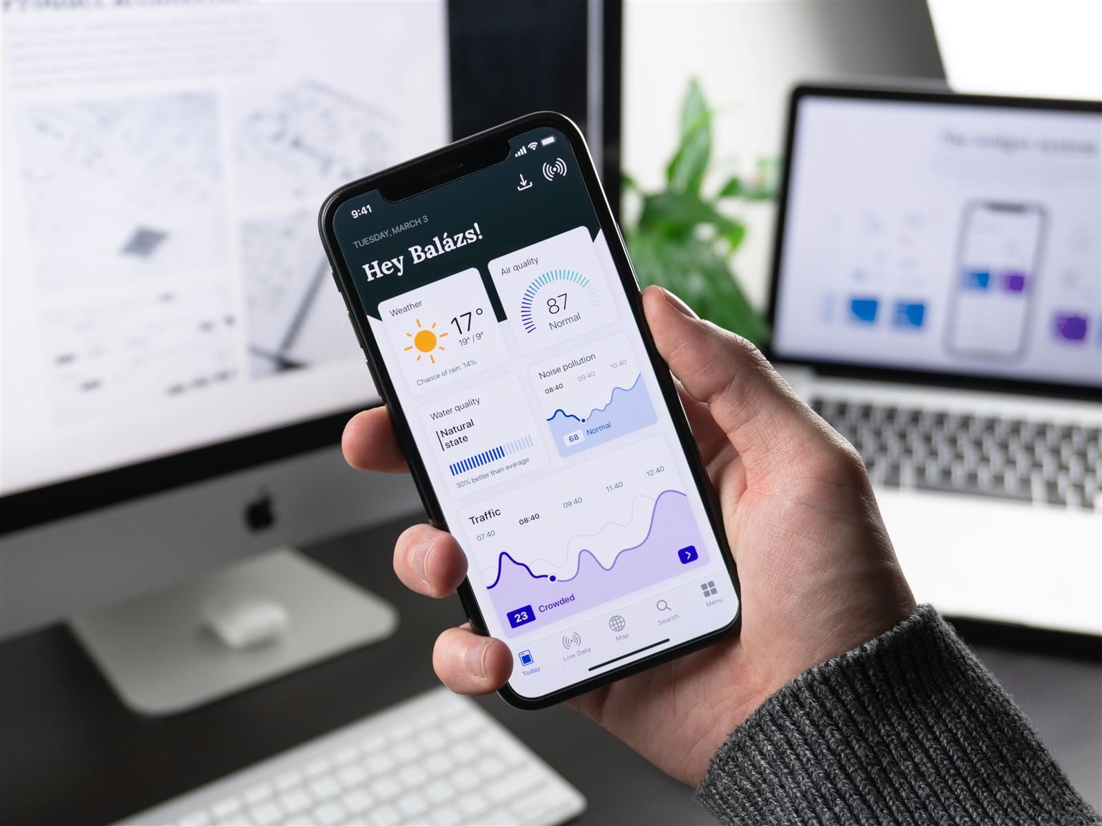 Hand holding a smartphone displaying a dashboard app, with a laptop and monitor visible in the background