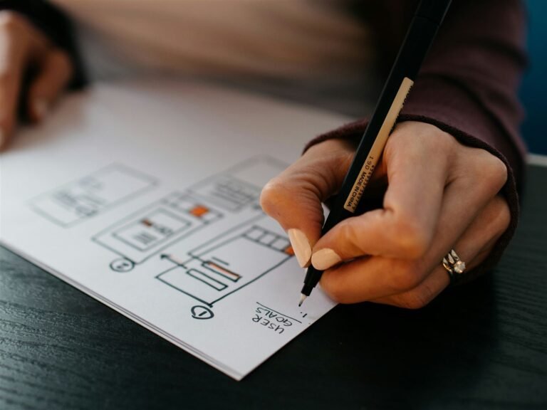 Hand sketching app screen wireframes on paper with a pen, with "user goals" written below